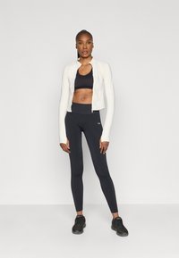 Λευκό ζακέτα με φερμουάρ, μαύρο αθλητικό σουτιέν και μαύρη κολάν. Λείο ύφασμα με εφαρμοστό σχέδιο, διακριτικό λογότυπο στα κολάν και αθλητικά sneakers.