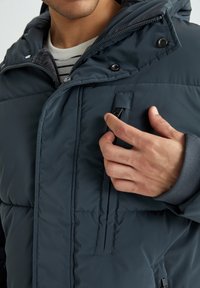 Mørkeblå vinterjakke med låsbar brystlomme, justerbar hætte og ribbede manchetter, med kviltet tekstur og glat overflade.