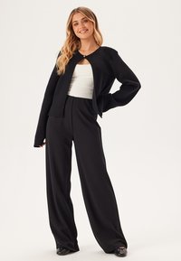 Svart öppen cardigan med lång ärm, tillsammans med vida svarta byxor. Cardiganen har en rundad halsringning och en minimalistisk design.