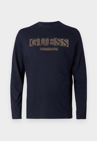 Tumesinise pikkade varrukatega T-särk, mis on valmistatud puuvillast, ja sellel on tekstuuriga kuldne "GUESS" logo rinnal ning ümmargune kaelus.