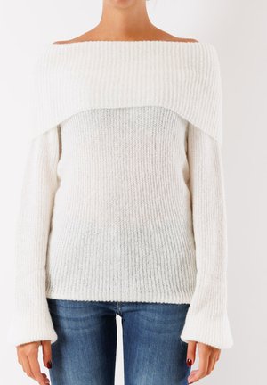 Maglione bianco a spalle scoperte in maglia con texture a costine, scollatura ampia e maniche svasate. Abbinato a jeans in denim blu.