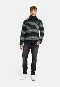 Grøn og grå stribet jakke med knaplukning, to brystlommer, båret over en sort højhalset top, parret med mørke jeans og hvide sneakers.