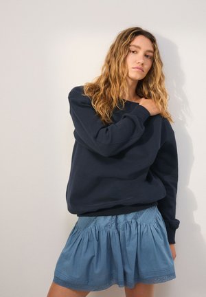 Kvinde med langt bølget hår står mod en ensfarvet væg iført en mørkeblå sweatshirt og en lyseblå plisseret nederdel.