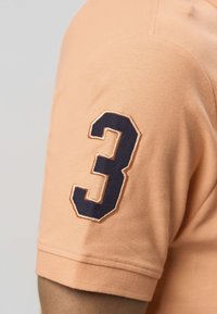 Narancssárga galléros póló texturált anyaggal, rövid ujjú, bal ujjon tengerészkék hímzett "3" számmal.