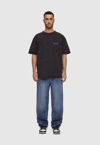 Svart oversized t-skjorte med lilla tekst på venstre bryst, sammen med løse blå jeans og svart-hvite sneakers.