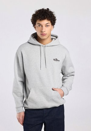 Giovane uomo con capelli ricci che indossa una felpa con cappuccio grigio chiaro con il logo "The Hamptons", mani nella tasca frontale, in piedi davanti a uno sfondo semplice.