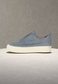 Ανεπίσημο παπούτσι τύπου slip-on με επάνω μέρος από ανοιχτό μπλε τζιν ύφασμα και παχύ λευκό λαστιχένιο πέλμα, πλευρική όψη πάνω σε ουδέτερη επιφάνεια και φόντο.