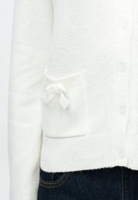 Fehér kötött kardigán texturált anyagból, gombos záródással és egy zsebbel, melyet egy kis fehér masni díszít.