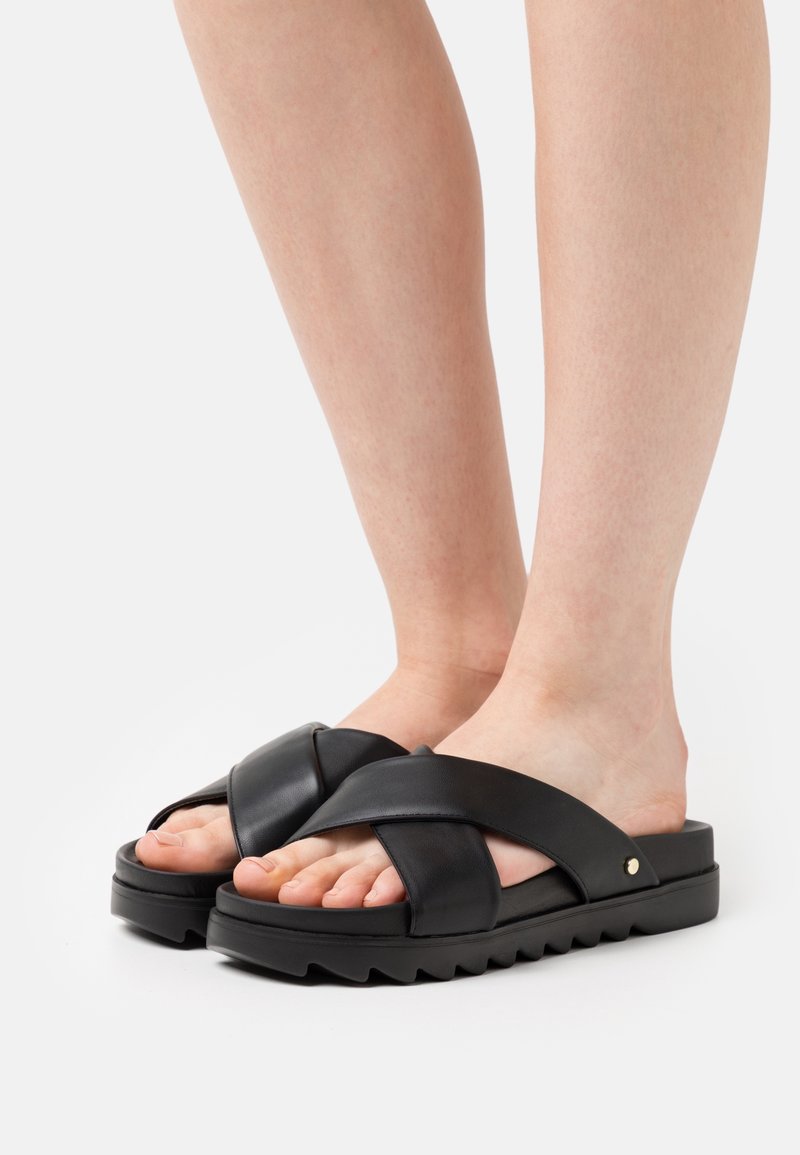 Sandales slide en cuir noir avec brides croisées, semelle épaisse et semelle inférieure texturée. Design à bout ouvert laissant la peau visible. Détail accent rond en or.