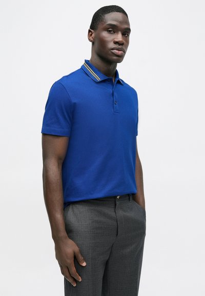 Karališkai mėlynas polo marškinėlis su dviejų mygtukų plokštele ir kontrastiniu geltonu dryžuotu apykakliu. Dėvimas su pilkais pritaikytais kelnėmis.