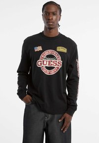 Zwarte t-shirt met lange mouwen, met op de voorkant en de mouwen graphics in rood en geel, voorzien van "GUESS" branding en een patch met een Amerikaanse vlag.