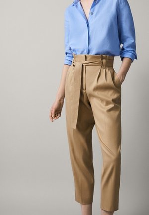 Kobieta ubrana w jasnoniebieską koszulę na guziki z podwiniętymi rękawami oraz wysokie spodnie paperbag karmelowego koloru z przewiązanym paskiem.