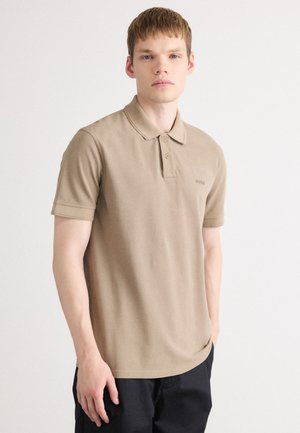 Jeune homme portant un polo beige avec deux boutons et un pantalon noir, se tenant devant un fond blanc uni.