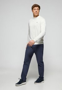 Maglione bianco a girocollo, pantaloni blu navy e sneaker scure. Il maglione ha maniche a coste e un piccolo logo. Texture del tessuto liscia e casual.
