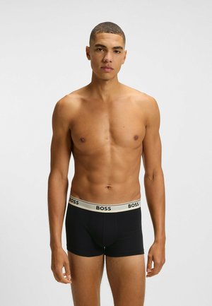 Ung man med kort hår som bär svarta BOSS-boxershorts med beige midjeband, stående mot en enkel ljus bakgrund.