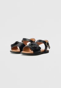 Sorte lær sandaler med ankelrem, med blomsteraksent foran. Flat såle, åpen design og glatt tekstur.
