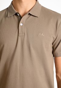 Brun polo skjorta i texturerad tyg, med krage, korta ärmar, två knappar och en liten bergslogotyp broderad på bröstet.