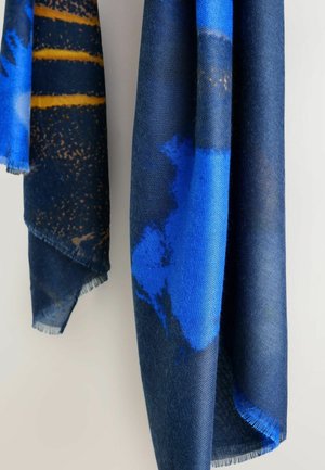 Austa skara su mėlyna ir tamsiai mėlyna abstrakčia raštais, su nuskilusiais kraštais ir minkšta tekstūra, kabančia vertikaliai prieš šviesų foną.