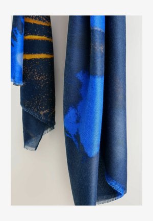 Kudottu huivi, jossa sininen ja laivastonsininen abstrakti kuvio, kuluneet reunat ja pehmeä pinta, roikkuu pystysuorassa vaaleaa taustaa vasten.