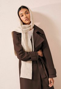 Brązowy płaszcz trench związywany w talii i szerokimi rękawami, zestawiony z jasnym, prążkowanym szalem. Gładka tekstura na tle jasnej ściany.