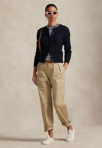 Marineblå kabelstrikket cardigan over en stripete skjorte, kombinert med beige plisserte bukser og hvite joggesko. Tilbehør inkluderer kattøyne-solbriller og et belte.