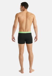 Černé plavky s zeleným páskem v pase, navržené pro těsné přilnutí. Látka působí hladce a pružně, vhodná pro vodní aktivity.