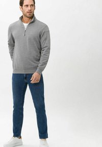 Pullover grigio con zip al collo realizzato in tessuto morbido, abbinato a jeans blu scuro e scarpe bianche, in piedi su uno sfondo chiaro.