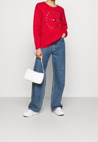 Persoon in een rode Tommy Hilfiger sweatshirt, blauwe wijde jeans, witte sneakers, die een kleine witte handtas vasthoudt tegen een effen witte achtergrond.