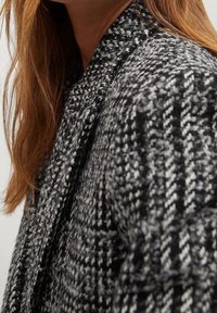 Manteau en laine à carreaux noir et blanc avec une surface texturée et un col châle, présentant une coupe décontractée et des détails de couture visibles.