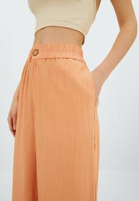 Leveälahkeiset housut pehmeästä, rypytetystä persikanvärisestä kankaasta. Niissä on korkea, joustava vyötärönauha koristeellisella napilla ja sivutaskut.