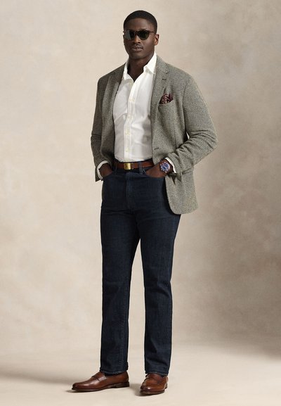 Mand iført solbriller, grå blazer, hvid skjorte, mørke jeans, brun bælte og sko, med hænderne i lommerne mod en neutral baggrund.