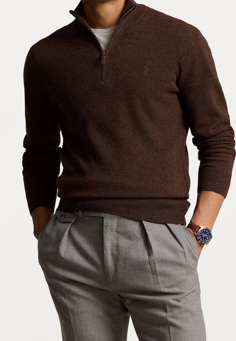 Homme portant un pull marron à fermeture éclair au quart, un pantalon plissé gris clair et une montre à bracelet en cuir marron avec un cadran bleu au poignet gauche.