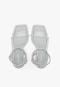 Transparante vierkante sandalen met verstelbare enkelsluitingen, voorzien van een glanzende afwerking en textuurdetails langs de zool in lichtgrijs.