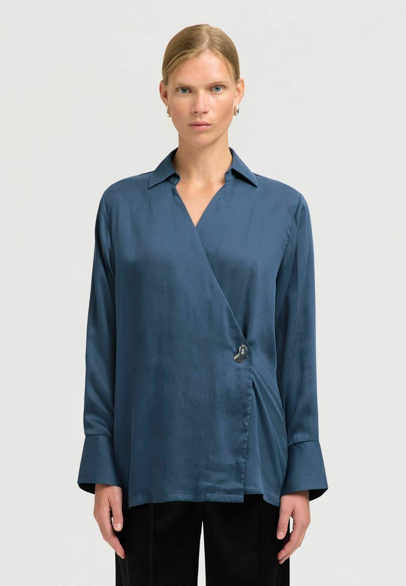 Camisa de satén azul con cuello puntiagudo, mangas largas y un gran detalle de botón decorativo en la cintura. Textura suave, corte elegante.