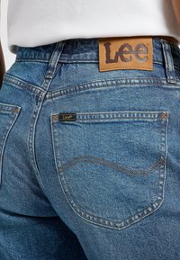 Jeans de mezclilla azul con una etiqueta de cuero de la marca Lee en la cintura, que presenta bolsillos traseros con detalles cosidos y un ligero desgastado.