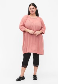 Tunika mekko lohenvärisessä, jossa on puffi hihat, pyöreä kaula-aukko ja rypytysdetaljeita. Mekkoa on yhdistetty mustiin polvimittaisiin legginsseihin, joissa on pitsireunus.