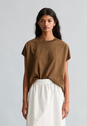 Vrouw met lang donker haar die een bruin kort mouwtshirt draagt, ingestopt in een witte rok met een hoge taille, poserend tegen een effen achtergrond.