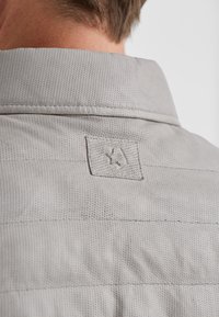 Hellgrauer Hemdkragen mit einer strukturierten Stoffoberfläche und einem kleinen, aufgestickten Logo-Patch. Der Kragen hat eine glatte Oberfläche und sichtbare Nähte.