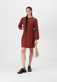 Rustfarget kjole med lange ermer, blonde detaljer ved halslinjen, belte i midjen og avslappet passform. Kombinert med svarte hæl-sandaler og en beige veske.