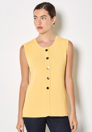 Kobieta z ciemnymi włosami w niskim kokie nosi żółty bezrękawnik z żebrowanej dzianiny z pięcioma złotymi guzikami z przodu oraz ciemne spodnie, patrząc w lewo.