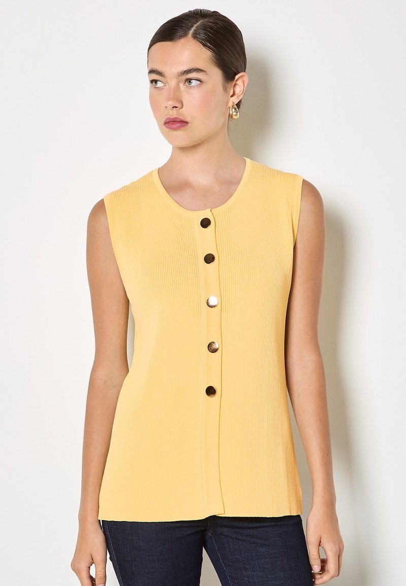 Femme aux cheveux foncés avec un chignon bas, portant un haut jaune sans manches côtelé avec cinq boutons dorés à l'avant et un pantalon foncé, regardant vers la gauche.
