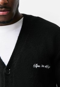 Zwarte gebreide cardigan met een diepe V-hals, voorzien van een enkele zwarte knoop en witte geborduurde tekst "Alpes Côte d’Azur" op de borst.