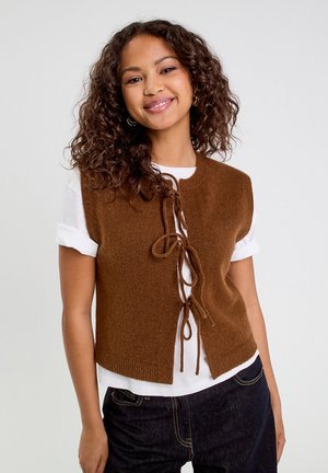 Bruine gebreide vest met een ronde halslijn en sluiting aan de voorkant met een strik, gedragen over een wit shirt met korte mouwen, gecombineerd met donkere jeans.