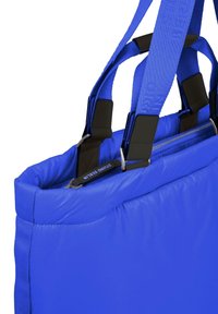 Blå vadderad axelväska med dubbla handtag, dragkedjestängning och kontrasterande svarta detaljer. Slät yta och strukturerad form.