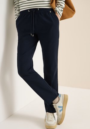 Personne portant un pantalon à cordon bleu marine, des baskets beiges avec des accents bleus, un t-shirt à manches longues rayé et un sac marron porté à l'épaule.