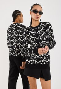Svart-hvitt mønstret genser med rund hals. Har lange ermer og ribbede mansjetter. Modellen har på seg svarte shorts; en annen modell er i bakgrunnen.