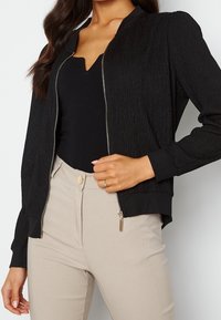 Giubbotto bomber nero con texture, chiusura a zip, polsini e orlo a coste. Abbinato a pantaloni beige a vita alta, con dettaglio di bottone frontale.
