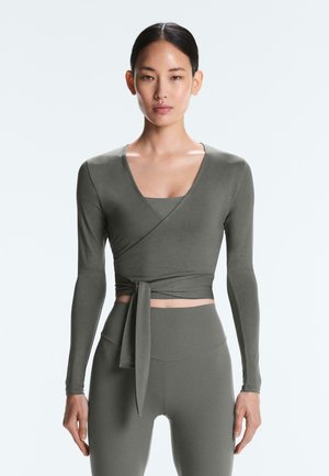 Frau trägt ein eng anliegendes, langärmeliges Wickeloberteil, das an der Taille gebunden ist, und hoch taillierte Leggings aus passendem, gedämpft grünem Stoff.