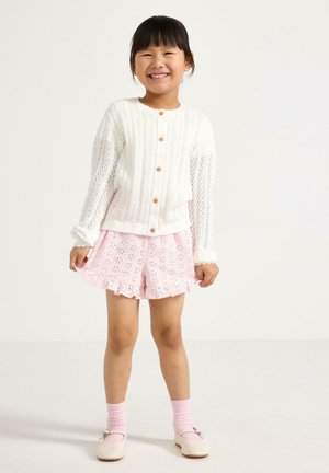 Smilende barn iført en hvid, knappet strikcardigan, lyserøde shorts med hulmønster, lyserøde sokker og cremefarvede sko med sløjfer, stående foran en ensfarvet baggrund.