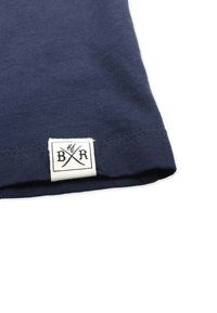 Navyblaues T-Shirt aus strukturiertem Baumwollmaterial, mit geripptem Halsausschnitt und einem weißen Stoffetikett mit schwarzem Logo und gekreuzten Linien.